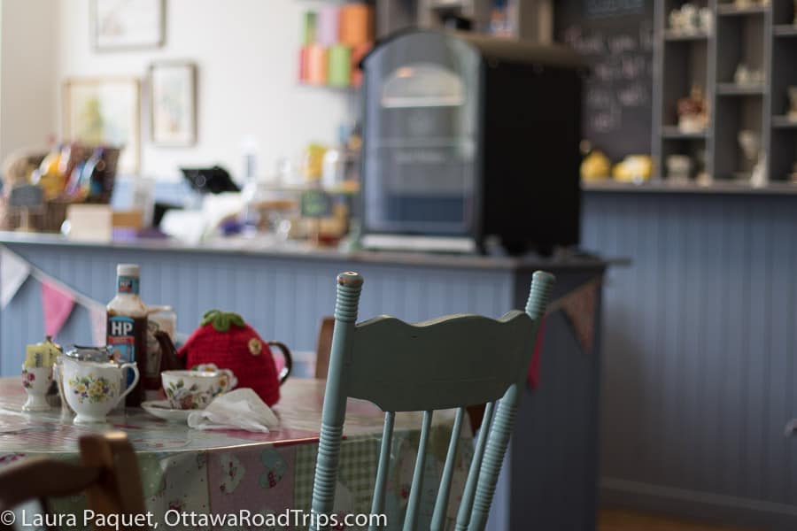 a blue chair at a table set with antique teacups and a brown betty teapot in a red knitted tea cozy, with cafe in background.