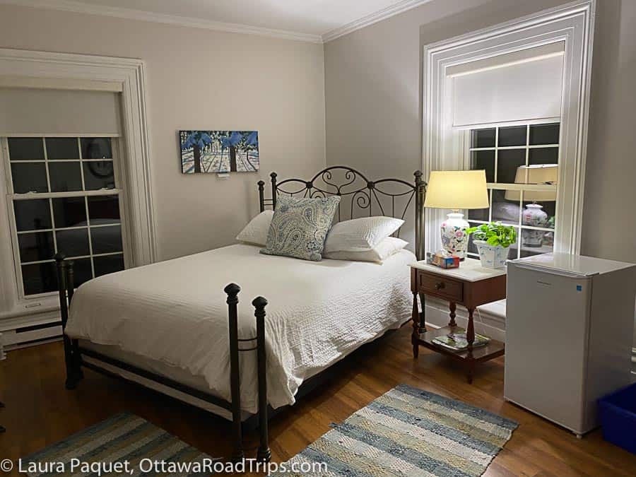 antique metal bed with white linens in a corner between two windows, in a guest room at the cove inn in westport, with hardwood floors, a small wooden nightstand and a mini-fridge.