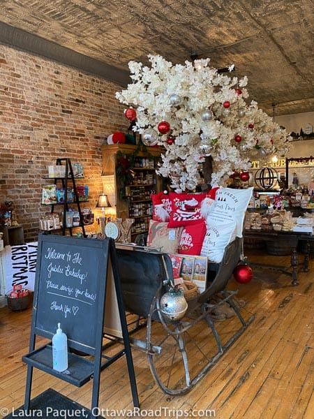 antique sleigh packed with christmas pillows; in the background, white tree with ornaments, wooden shelves with gifts and baked goods.