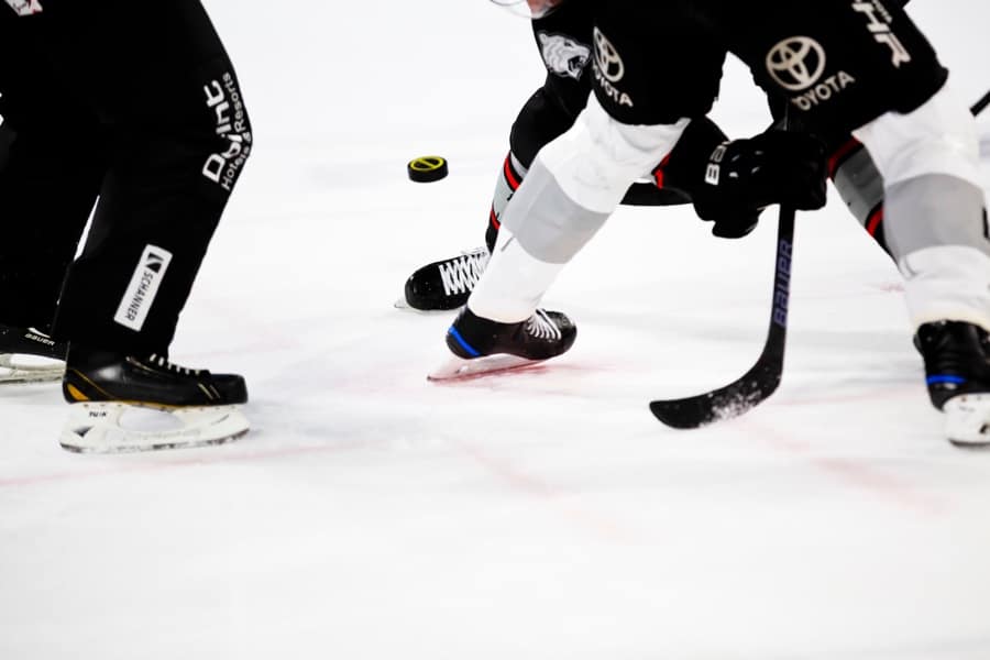 Closeup of several players' feet in skates on the ice, with a hockey stick in the foreground and the puck in the background.