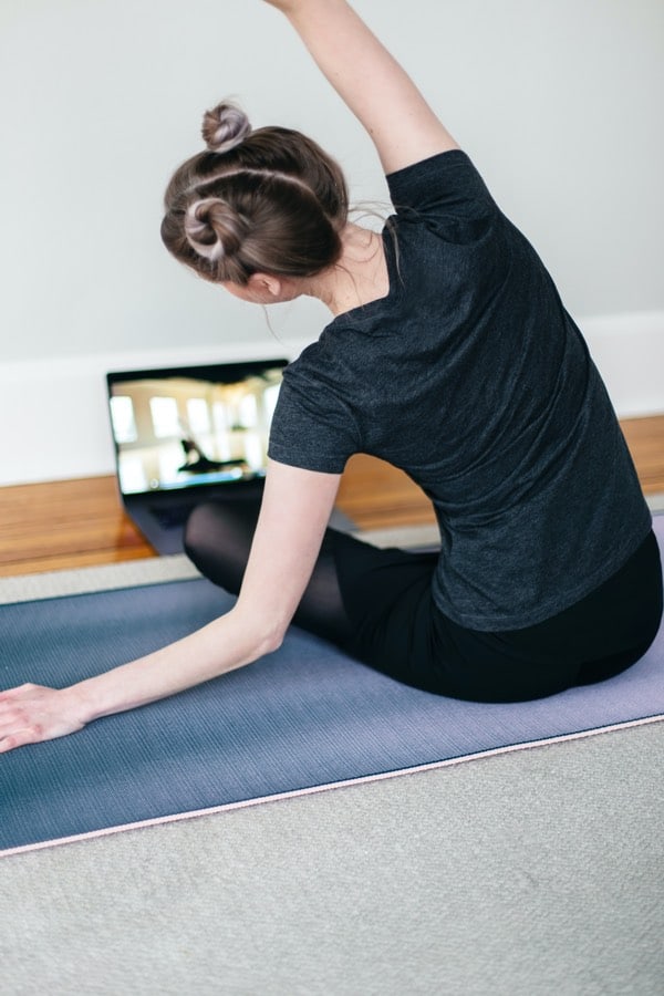 young woman in a grey t-shirt sitting cross legged on a yoga mat and stretching in front of a laptop