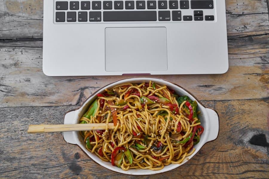 Bowl of noodles and vegetables in front of a laptop