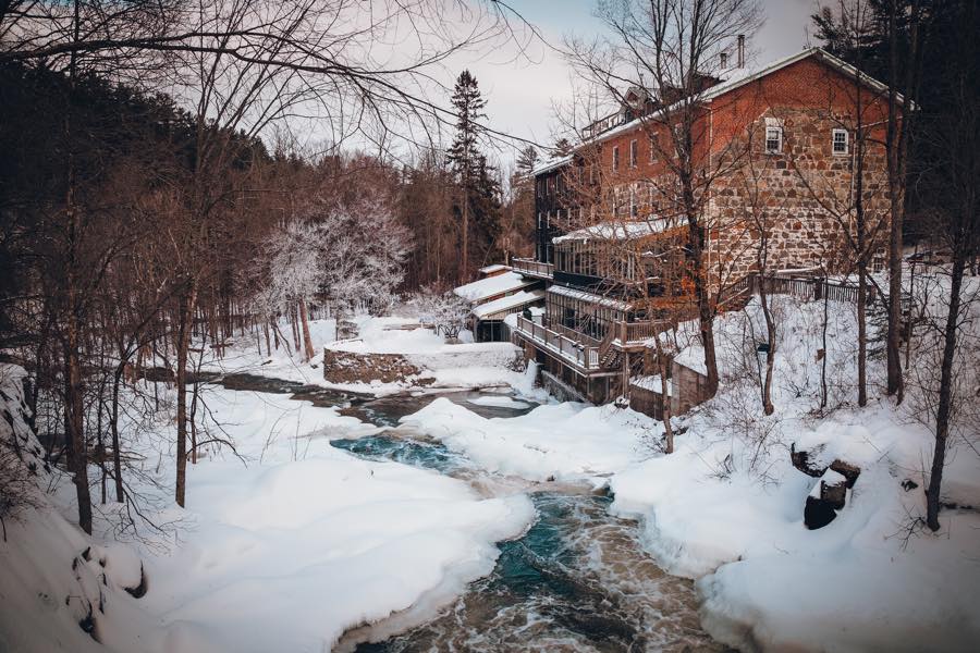 old stone mill with wooden deck overlooking frozen river and snowy trees.