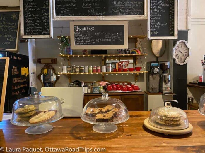 wooden counter with baked goods on glass stands under domes and chalkboards describing menu in background at bubba & bugs coffee bar in kemptville