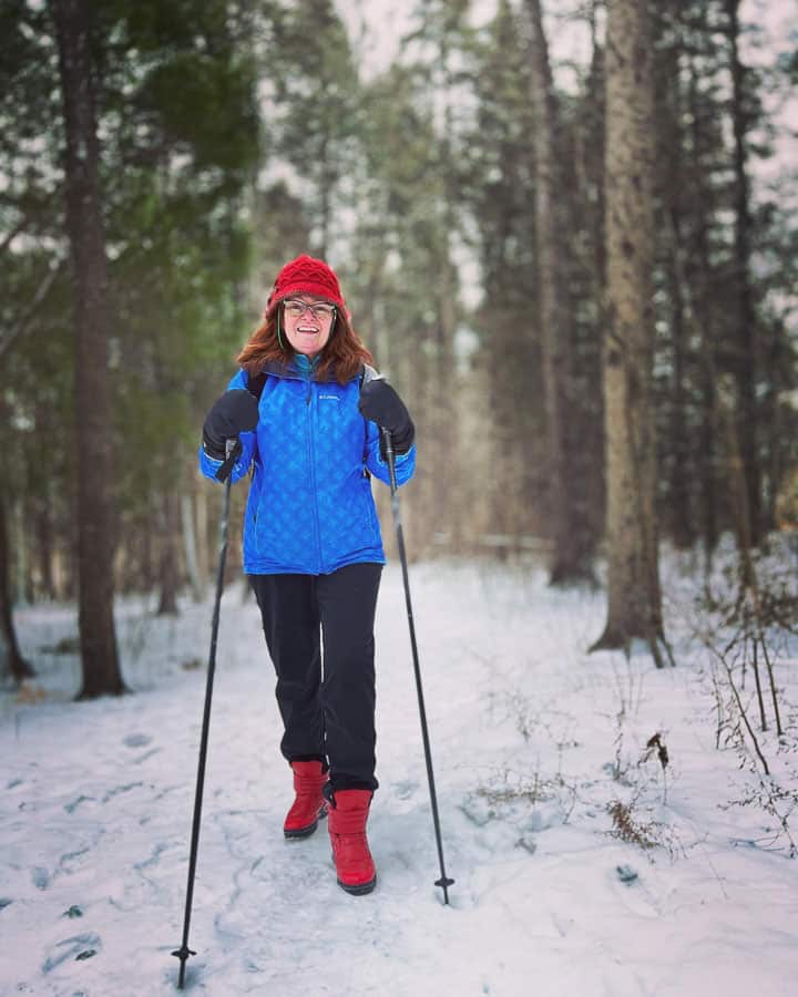 woman in blue jacket and red hat walking along winter trail with ski poles
