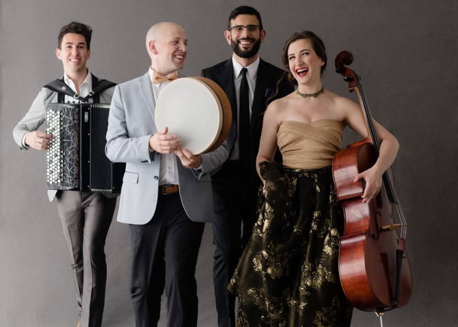 Three men and a woman in formal clothes, laughing and holding instruments, including an accordion and a cello