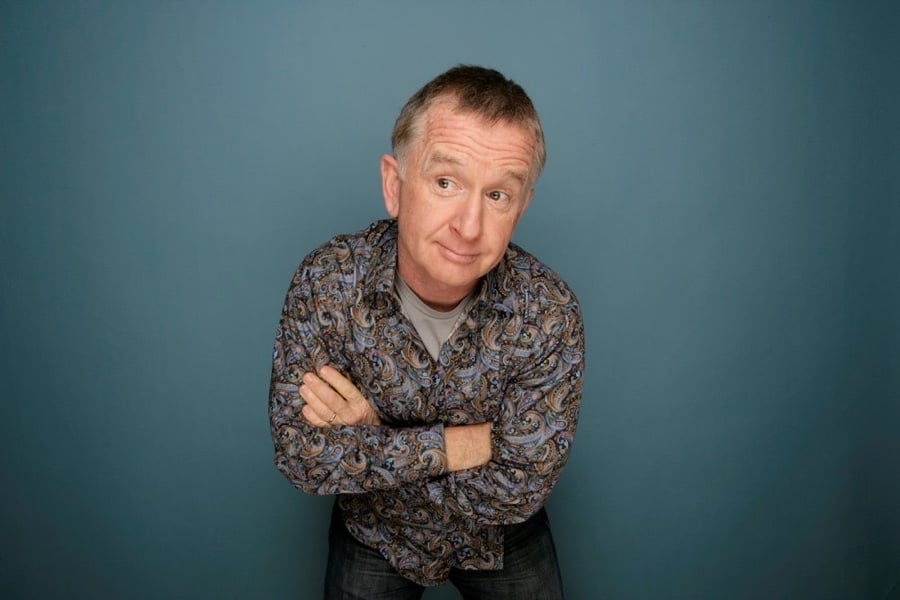 Man with greying hair standing with arms crossed against a blue background