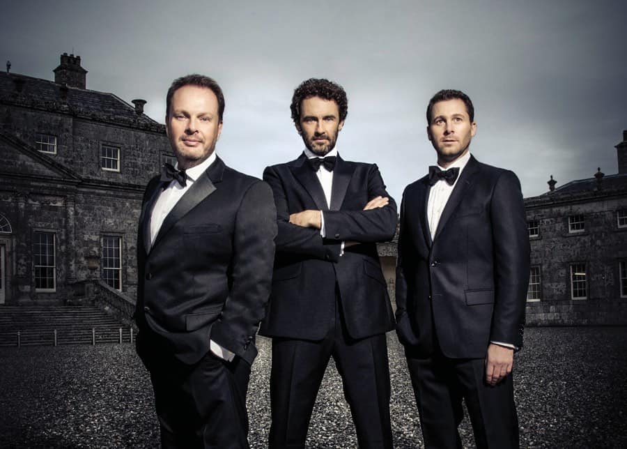 Three dark-haired men in tuxedos, standing in front of two old stone buildings with mullioned windows.