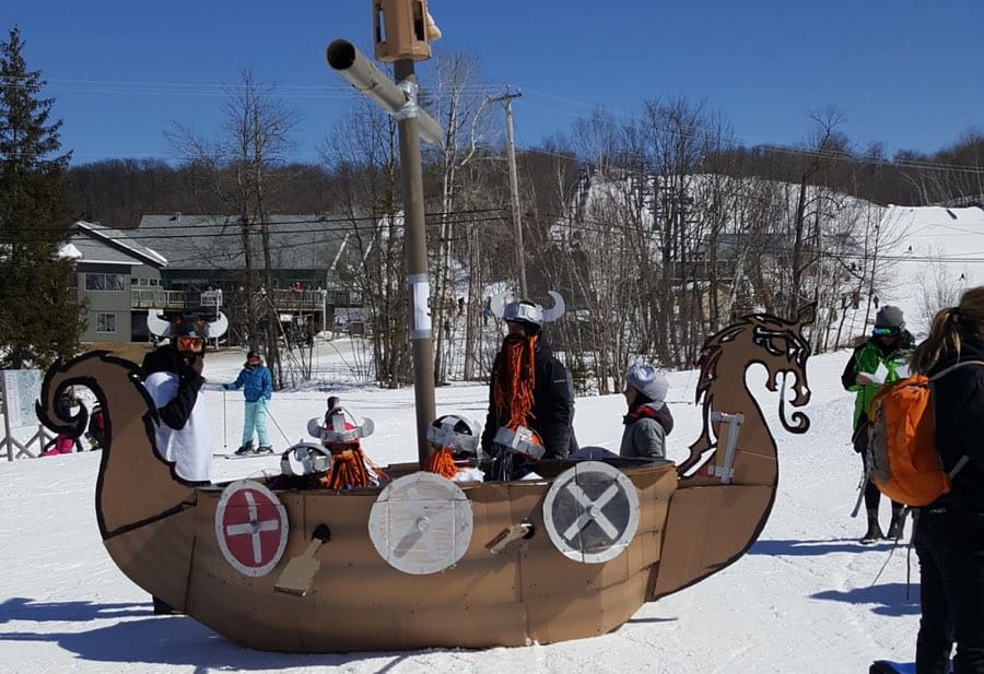 Kids and adults wearing Viking helmets inside a cardboard Viking ship on a snowy ski slope.