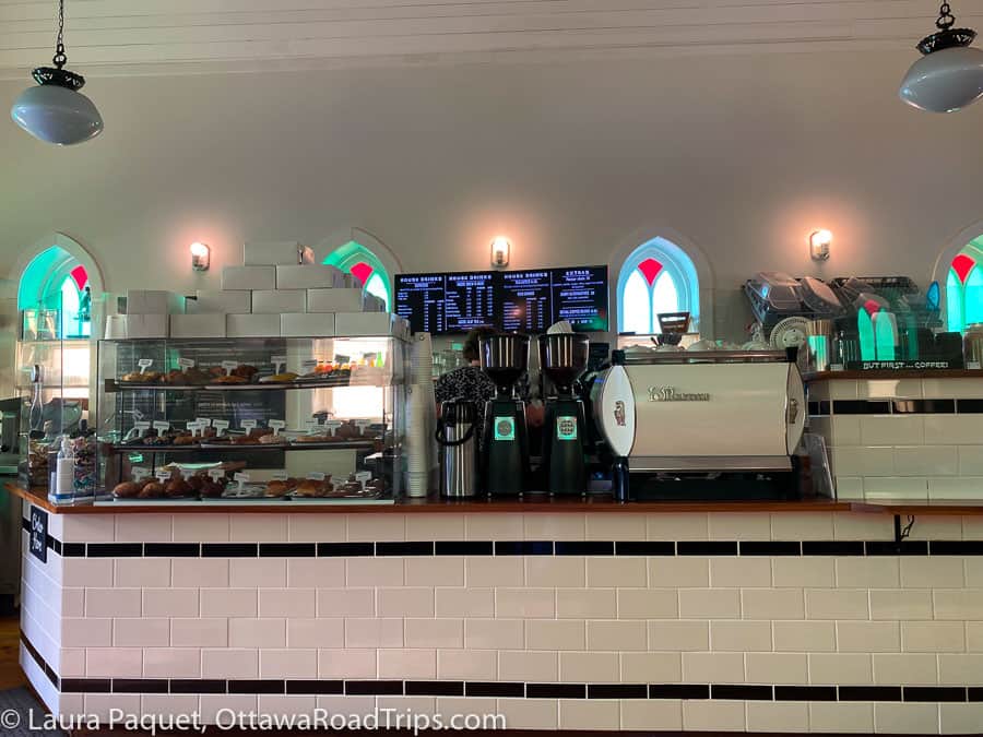 large café counter with black-and-white subway tiles and wooden top, with simple arched stained-glass windows in background, at the cardinal café + shop in sharbot lake, ontario.