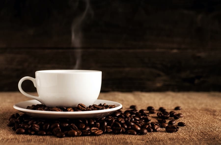 Small white cup on a saucer, with roasted coffee beans on the saucer and the table around it.