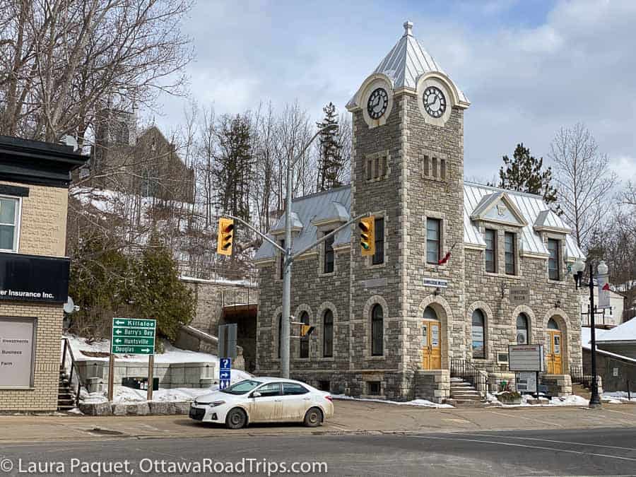 the bonnechere museum in eganville, ontario, is a limestone building with arched windows, a silver roof and a clock tower.