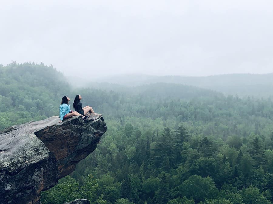 two women sitting on the eagle's Nest Lookout, a rocky outcrop overlooking green trees and misty hills in Calabogie, Ontario.