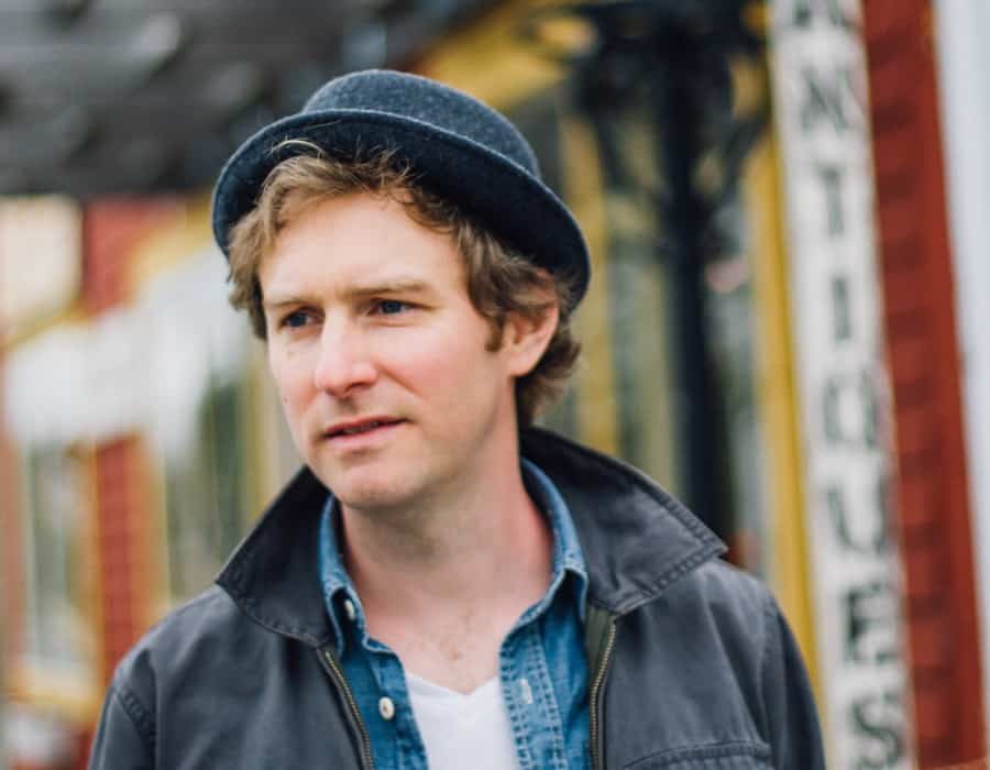 Man with curly light-brown hair wearing a grey brimmed hat in front of a blurred background.