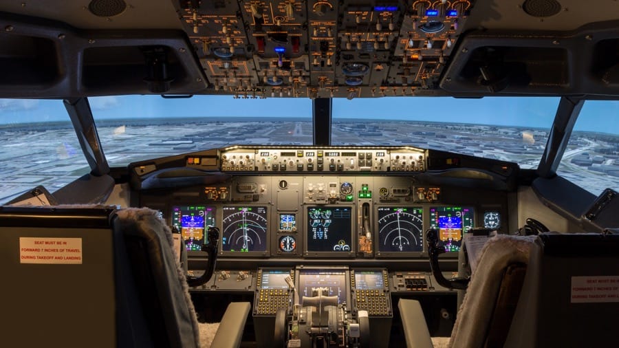 cockpit of an aircraft simulator with lighted controls and a simulated view of an airport through the windscreen.