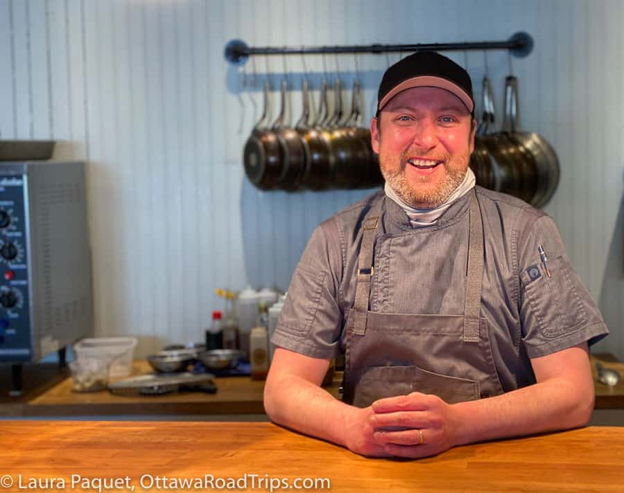 man in grey chef shirt and apron, with black baseball cap, smiling behind wooden counter with pots hanging in background, at the Axe & Paddle restaurant in Eganville, Ontario