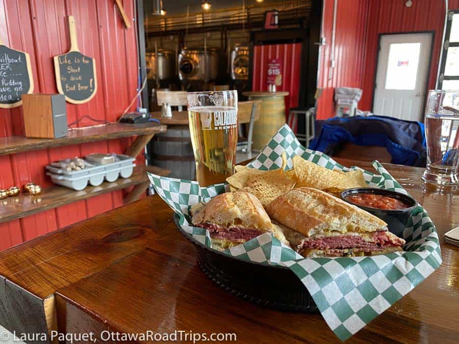 a large sandwich, with tortilla chips and salsa, and a pint of beer, on a wooden table with silver beer-brewing equipment in background at the calabogie brewing company.