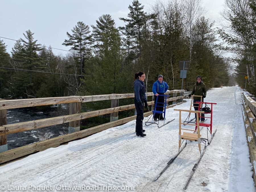 two women and a man standing with kicksleds on a snow-covered wooden bridge above the madawaska river in calabogie, ontario