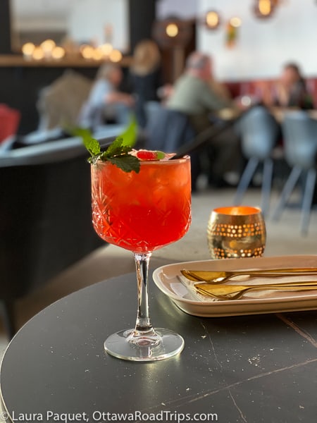 orange-red cocktail garnished with mint leaves in a large crystal wine goblet with blurred restaurant in background