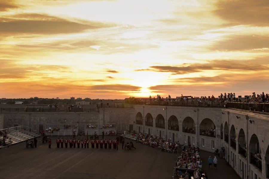 crowds in galleries and on top of limestone wall at fort henry in kingston, ontario, watching guards in red coats on drill field at sunset.
