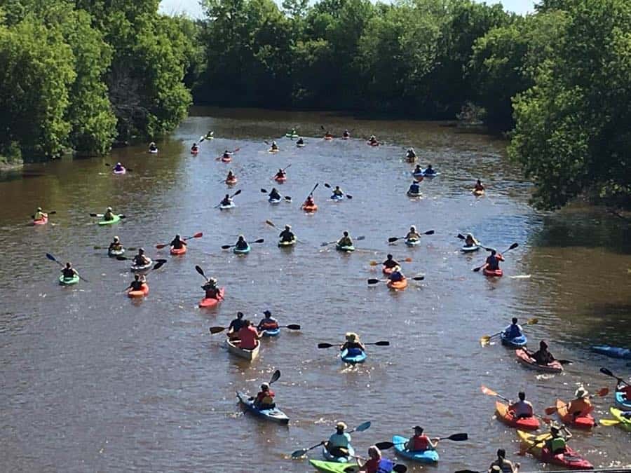 About 30 kayakers paddling down a river, away from the camera.