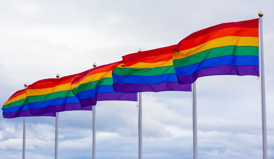 multiple rainbow pride flags on silver flagpoles against a cloudy sky