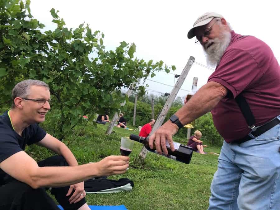 man pouring red wine into glass of man sitting on yoga mat, with vineyard and other yogis in background