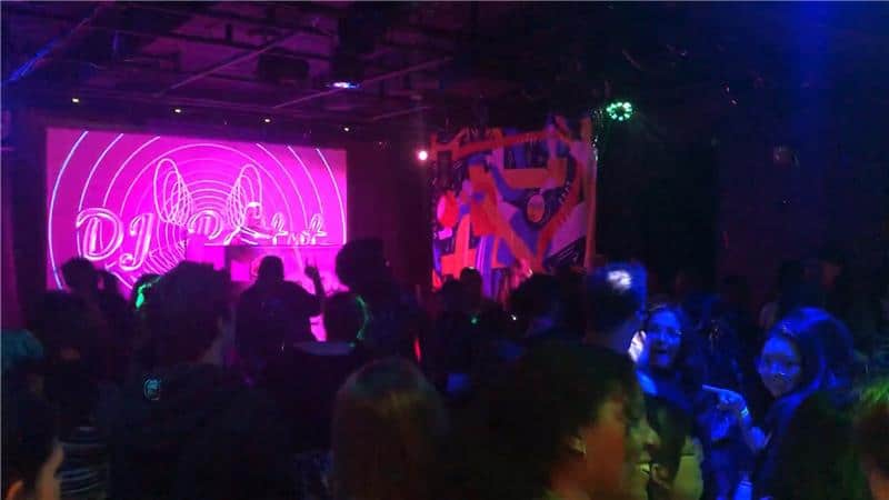 People dancing in a dark room lit by multicoloured lights and projections on screens.