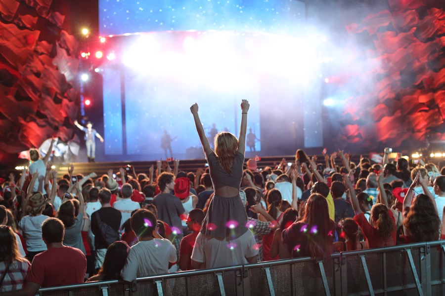 Fans waving arms in the air at a rock concert, photographed from behind, with stage in the distance.