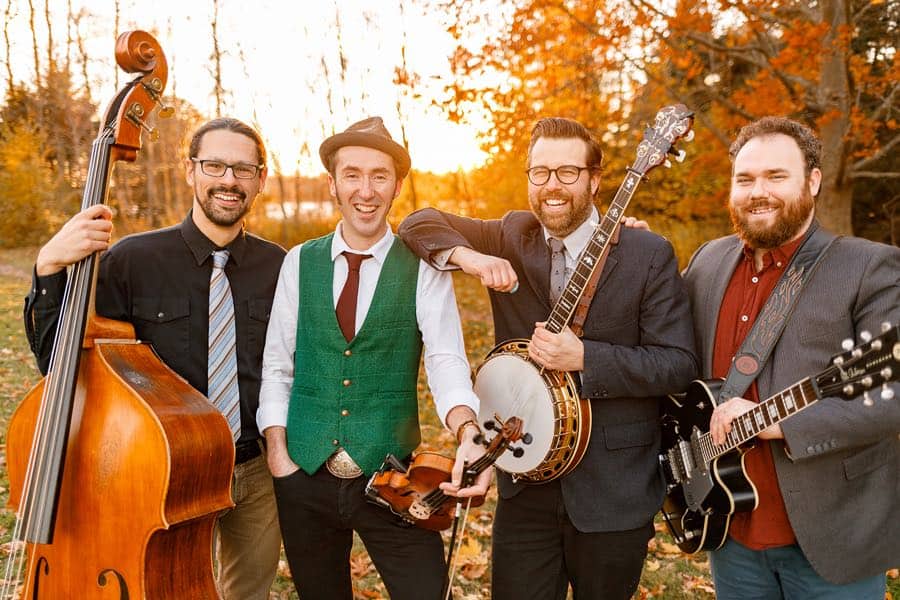 Four men in jackets and ties holding stringed instruments and standing in a forest in fall.