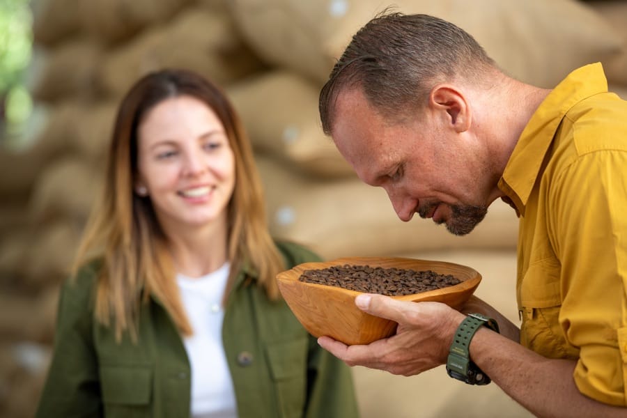 Man sniffing coffee beans in a wooden bowl while woman watches in background