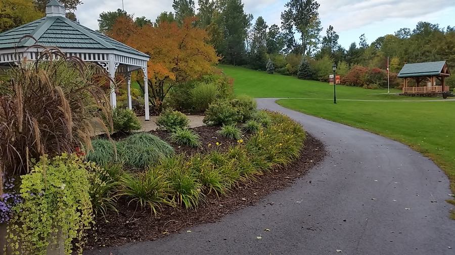 curving paved path across a green lawn, with gardens, trees, a gazebo and a picnic shelter