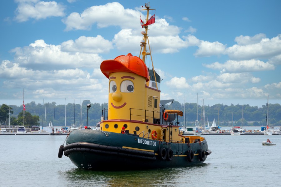 Theodore Too (large yellow tugboat with a smiling face) in a harbour with sailboats and dock in background