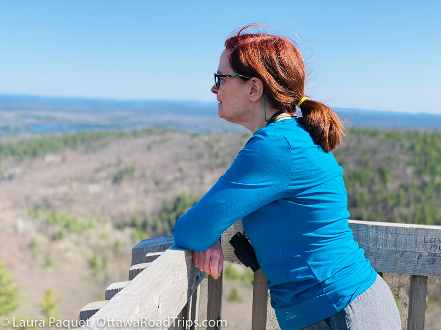 woman with glasses and a ponytail looking over wooden railing at landscape of lakes and trees