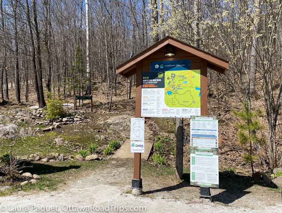 map and other signs under a small wooden roof, with trail and trees behind, at the mont morisette trailhead.