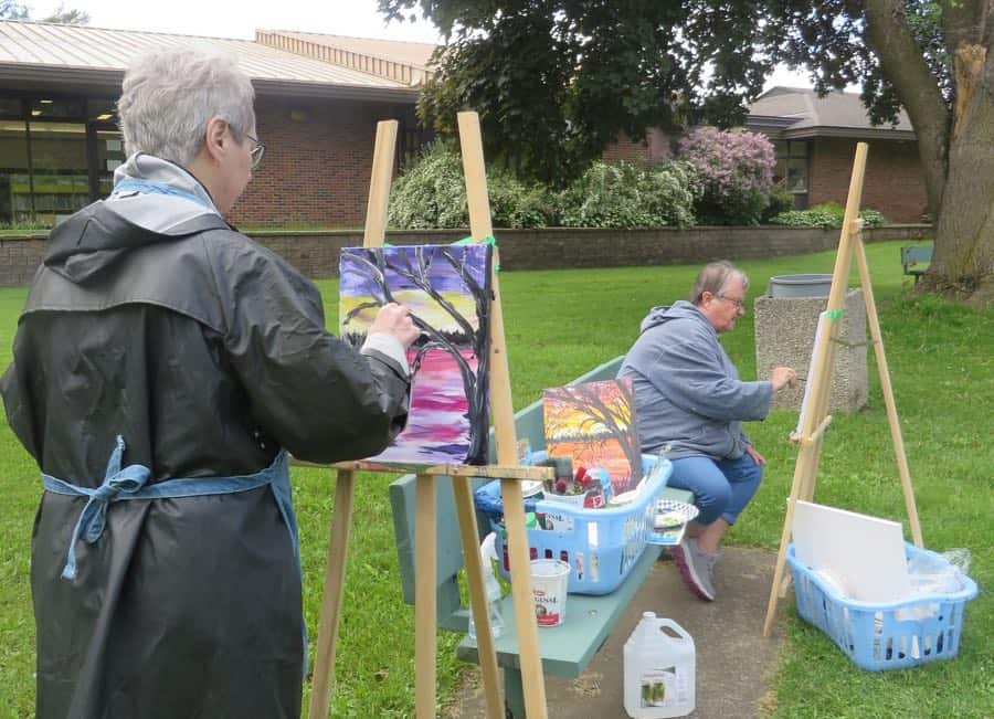 two women painting outdoors on easels, with red brick building in background