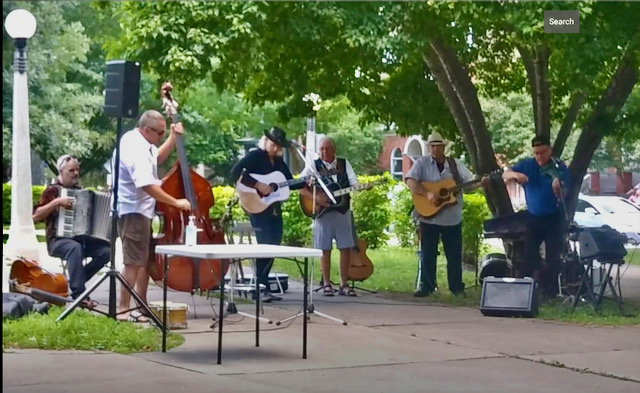 Six men playing various instruments (guitars, accordion, bass) outdoors under trees.