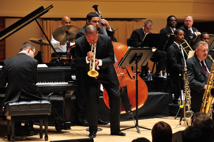 Wynton Marsalis leaning forward playing trumpet with orchestra in background in a concert hall.