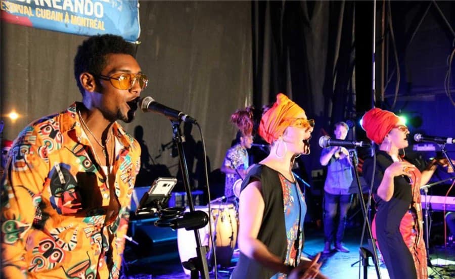 Two women and a man in colourful clothing singing at microphones, with musicians in background.