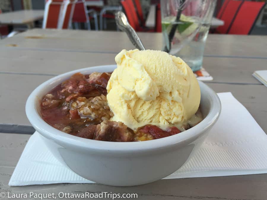 close up of a fruit crumble with a scoop of vanilla ice cream in a white bowl on a wooden picnic table