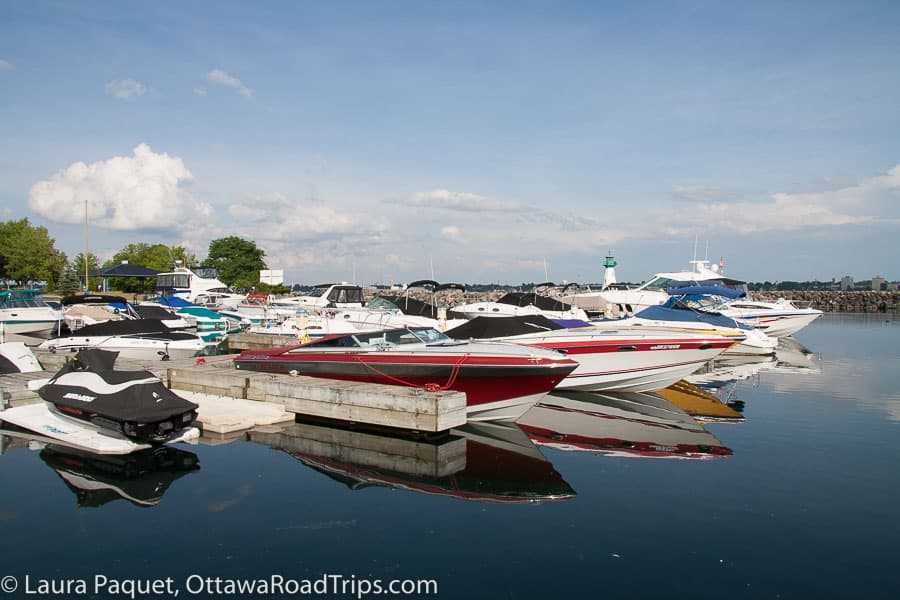 motorboats and speedboats at a marina, reflected in water with small green-and-white lighthouse in background at sandra s lawn marina in prescott, ontario.