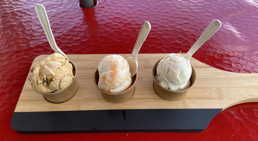 three small cups of ice cream with wooden spoons on a wooden cutting board atop a red table.