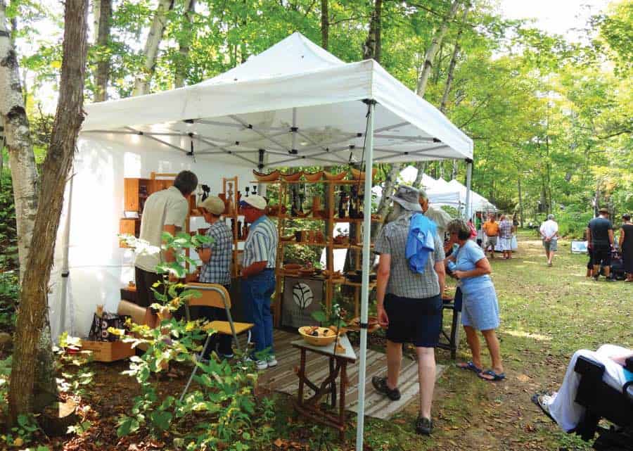 People in summer clothes browsing for items under white tents set up below trees.