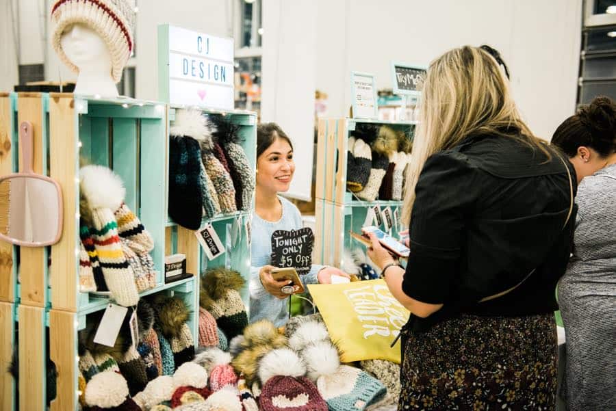 Smiling woman at a craft fair booth surrounded by knitted hats, with two customers.