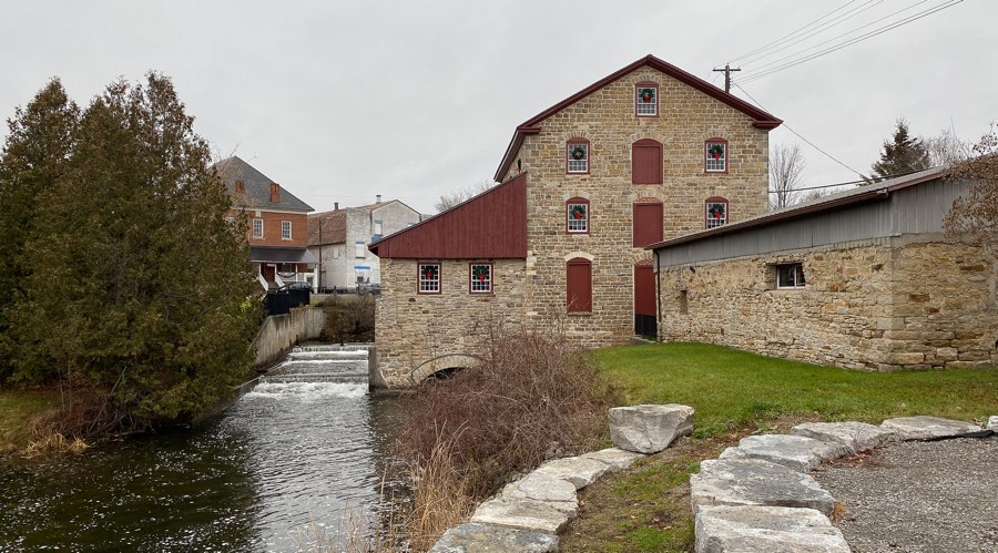 four-storey limestone old stone mill in delta, ontario, next to a watercourse, with houses in background.