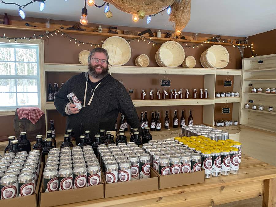 bearded man behind a large table holding lots of tins of apple cider at coronation hall cider mills.