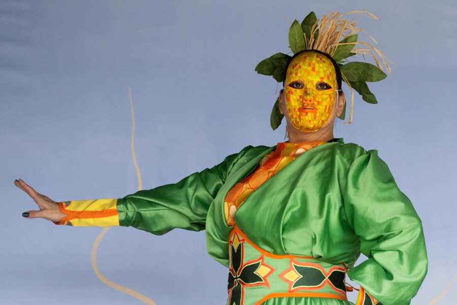 Person in elaborate green, orange and yellow robes with an orange and yellow face mask, and green leaves and grasses on their head.