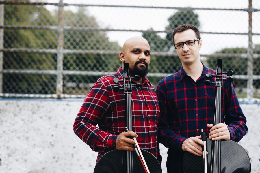 Two men in plaid shirts holding the necks of cellos, with a chain-link fence in the background.