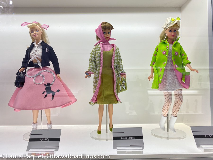 three dolls, one with a pink poodle skirt, one in a green dress with a 1950s-style head scarf and one in a white mini dress with green coat