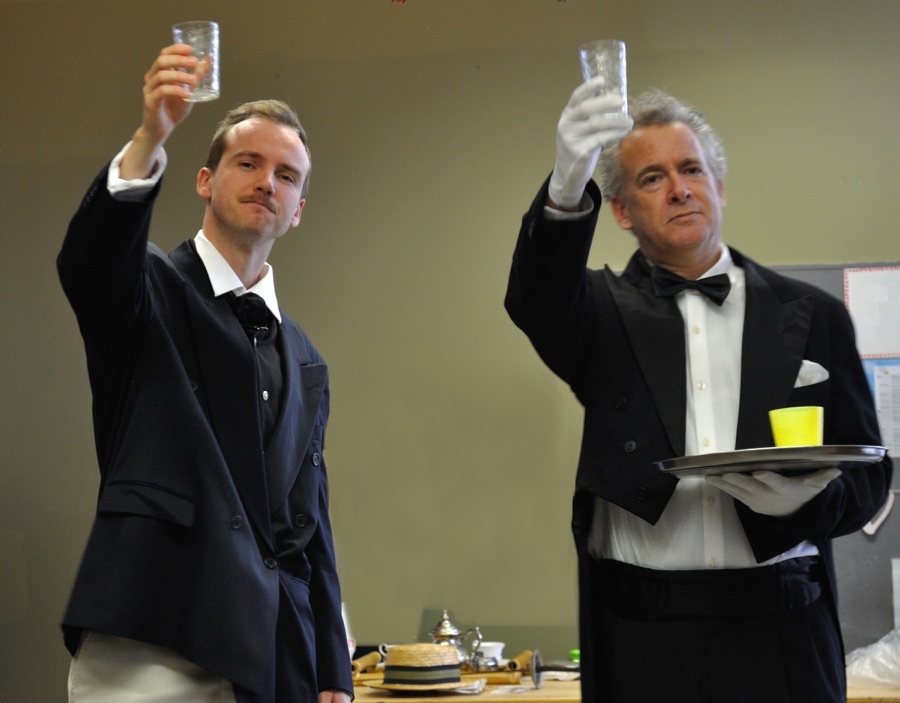 Two men in vintage evening wear, holding up glasses in a toast. The oder man is wearing gloves and carrying a silver tray.